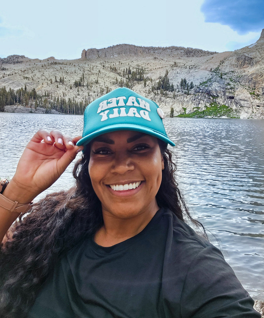teal hat with the words Water Daily on the hat on a woman with long curly hair looking with a mountain scene. 