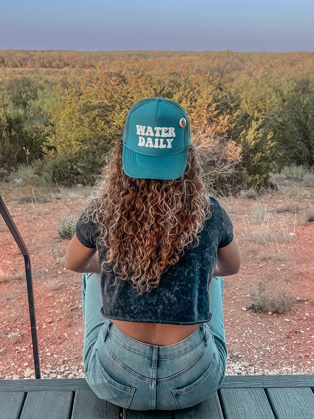 teal hat with the words Water Daily on the hat on a woman with long curly hair looking out at a desert scene. 