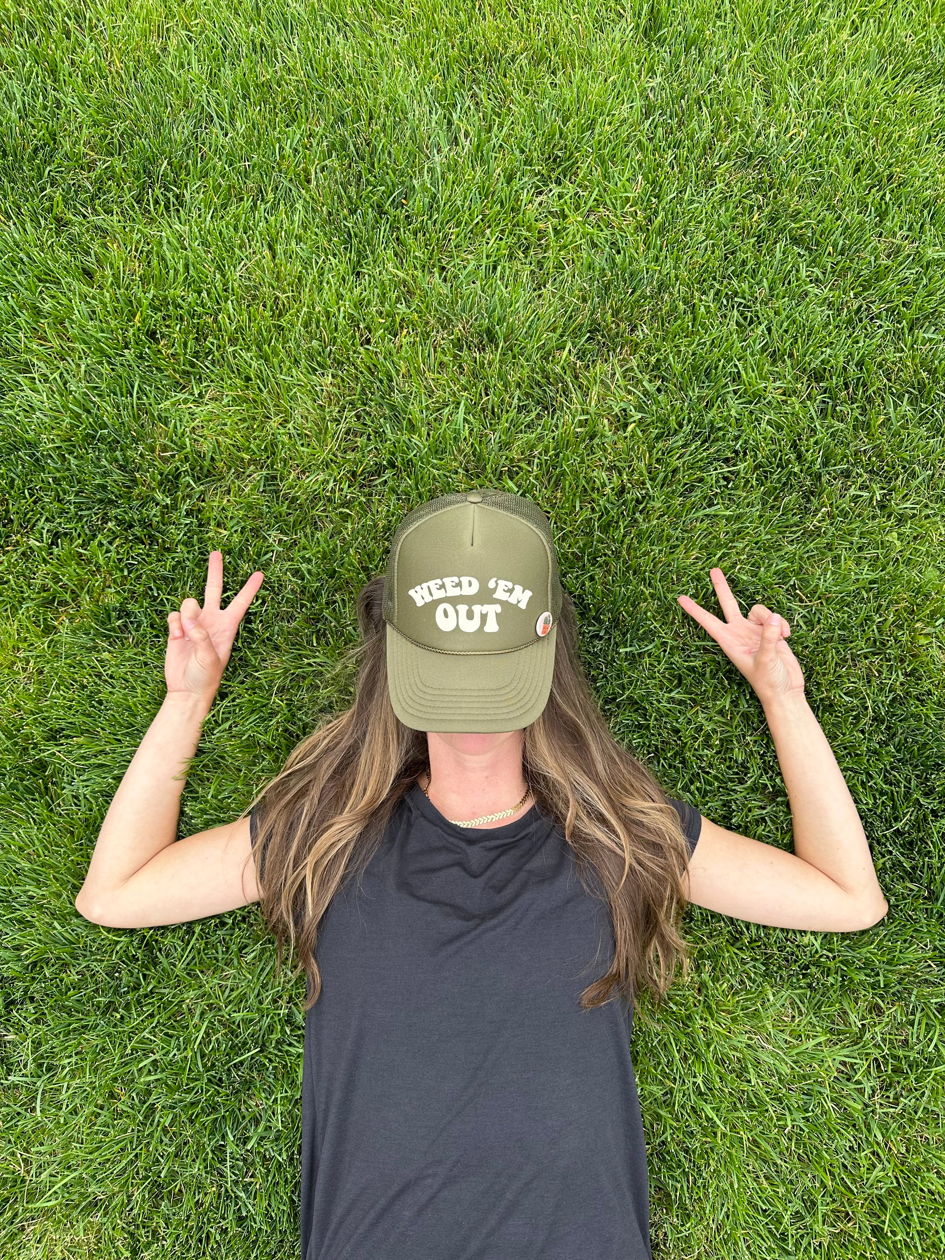 olive green hat with the words Weed Em' Out on the hat. Background behind hat with green grass and a woman with long hair is wearing the hat.