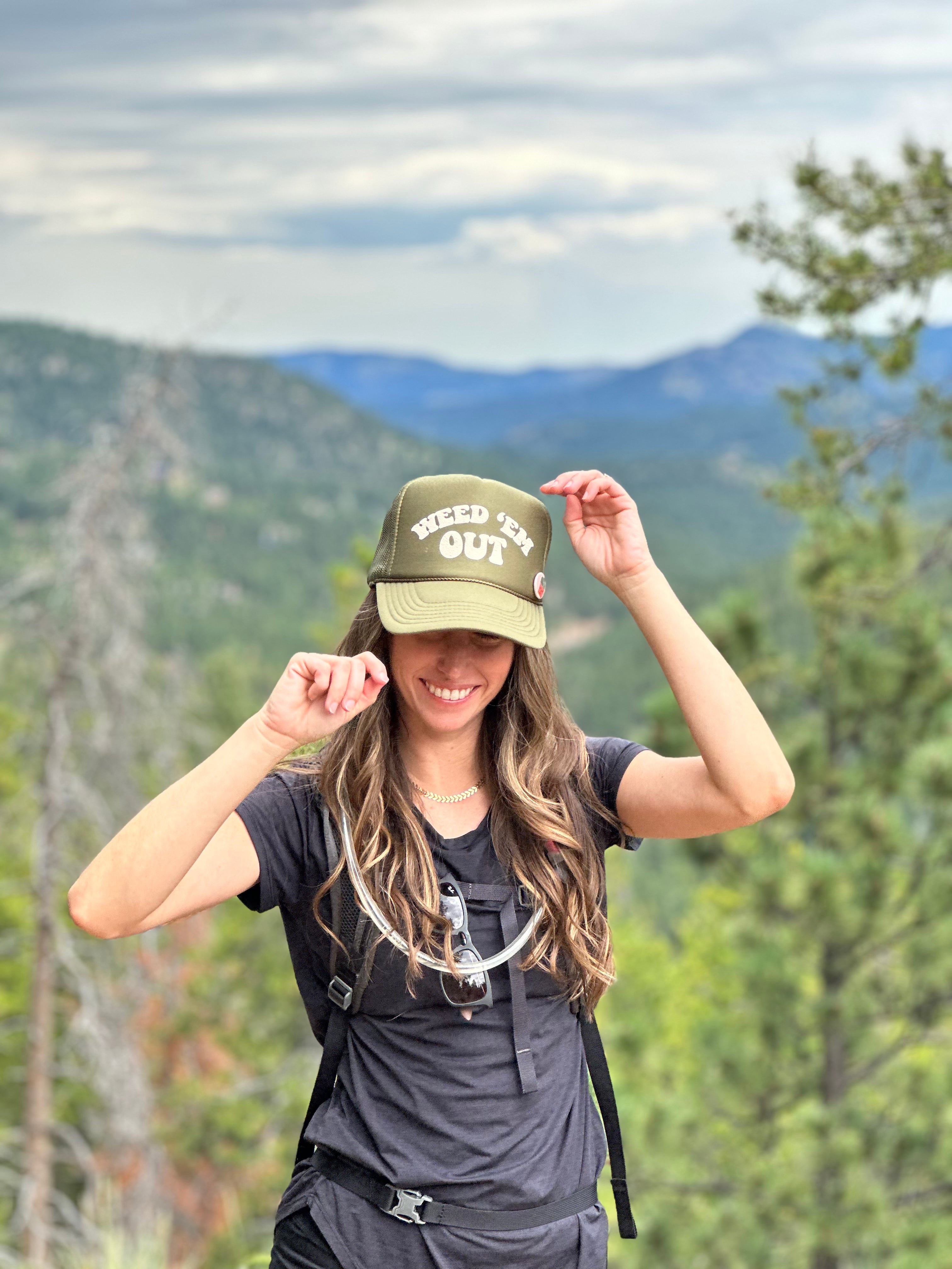 olive green hat with the words Weed Em' Out on the hat. Background behind hat is a mountain scene and a woman with long hair is wearing the hat.