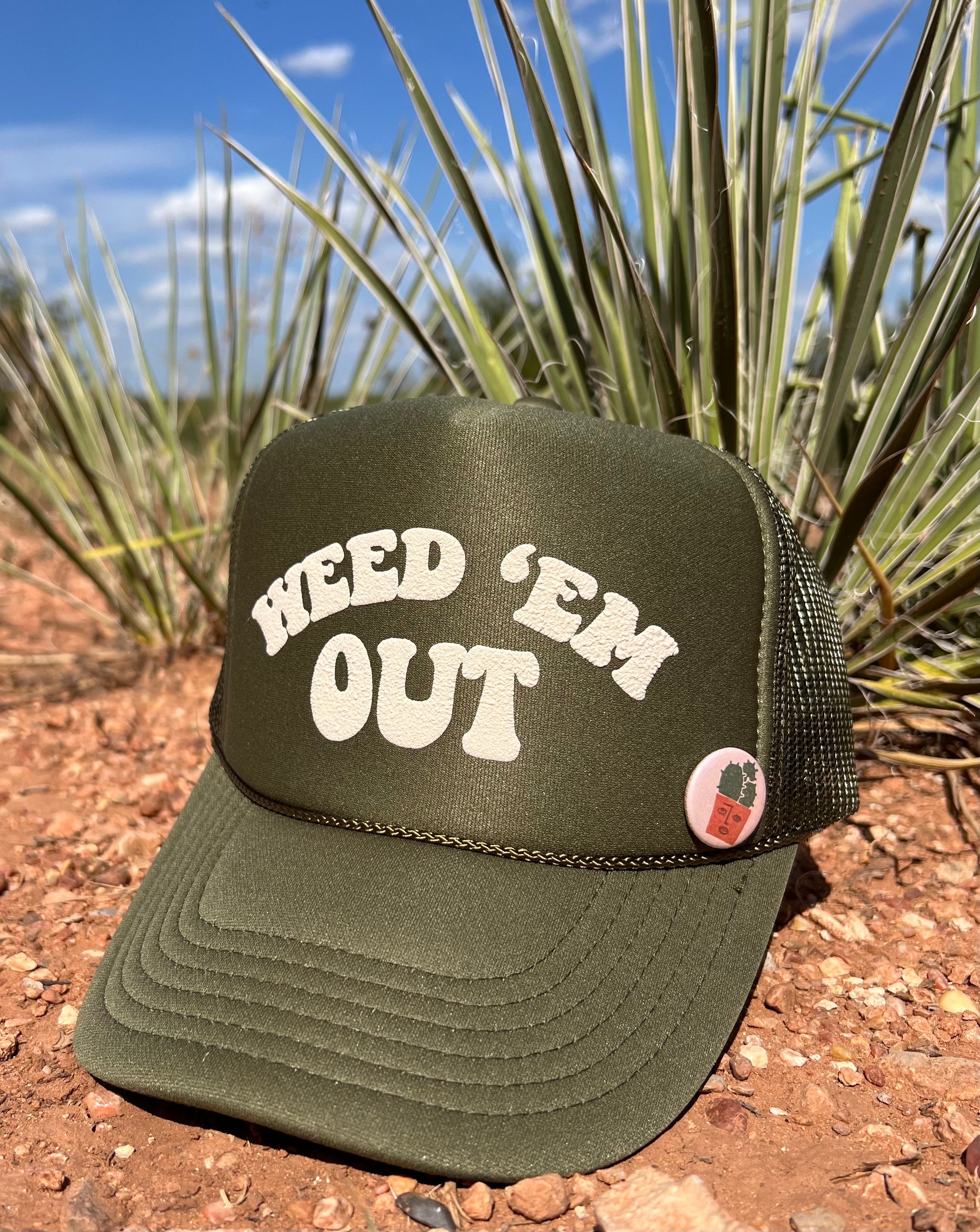 olive green hat with the words Weed Em' Out on the hat. Background behind hat is a desert.
