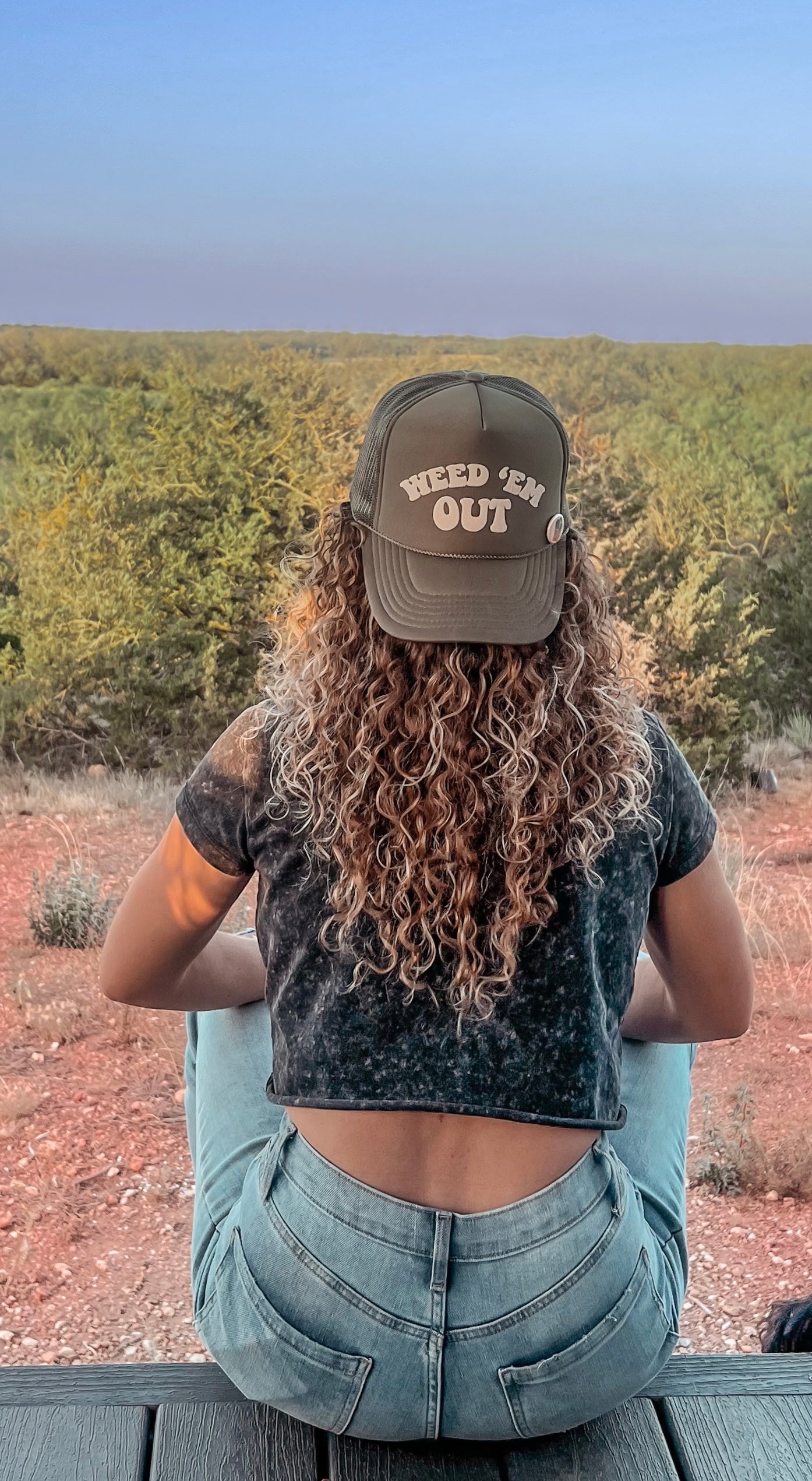 olive green hat with the words Weed Em' Out on the hat. Background behind hat is a desert and a woman with long curly hair is wearing the hat.