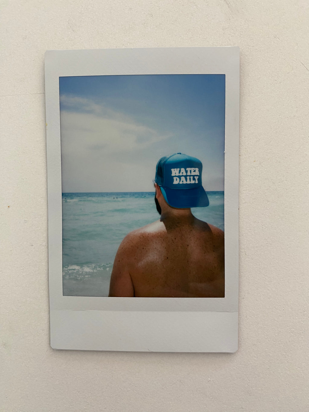 teal hat with the words Water Daily on the hat on a man with looking out at the ocean.