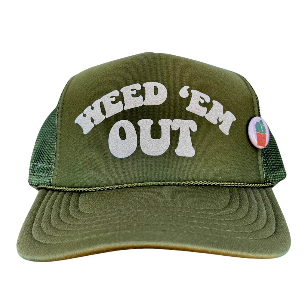An olive green trucker hat with the text 'WEED 'EM OUT' in white letters on the front and a small pin with a plant illustration on the left side.