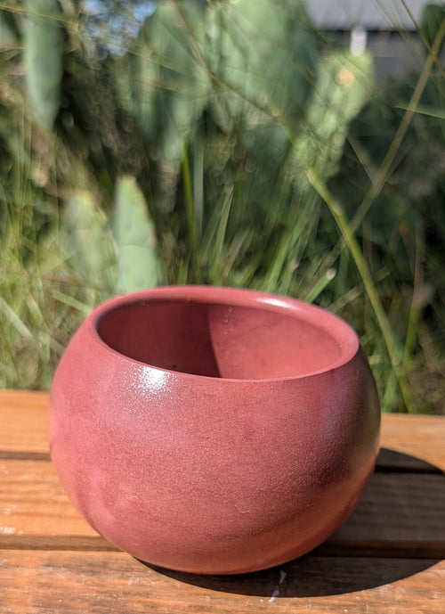 A small round terracotta-colored planter pot with a matte finish, sitting on a wooden deck table outdoors with green plants blurred in the background.